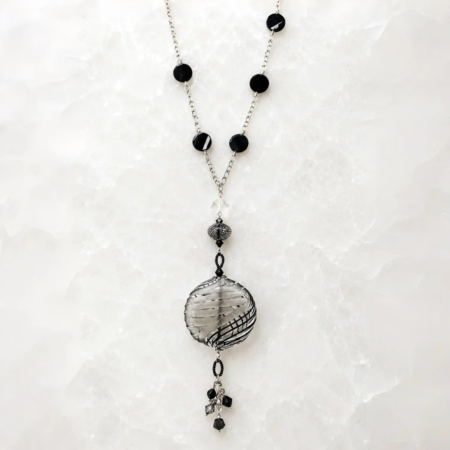 Venezia Murano glass necklace handcrafted in Canada with sterling silver chain, black Murano glass stations, artisan Murano pendant and crystal tassel on white quartz background.
