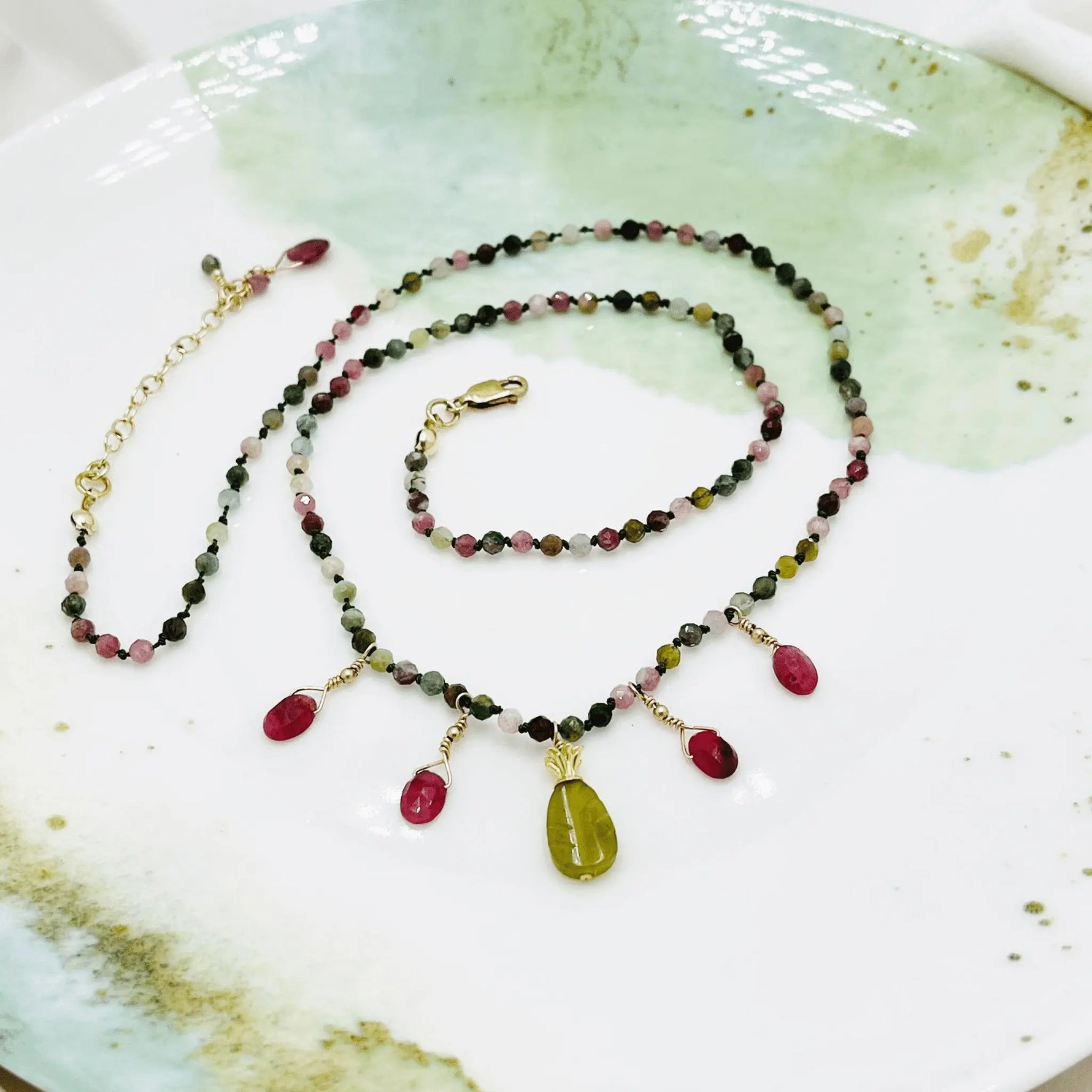 Styled flat lay of the Layla Gemstone Necklace with tourmaline beads, ruby accents, and a carved jade leaf pendant arranged on a ceramic dish.